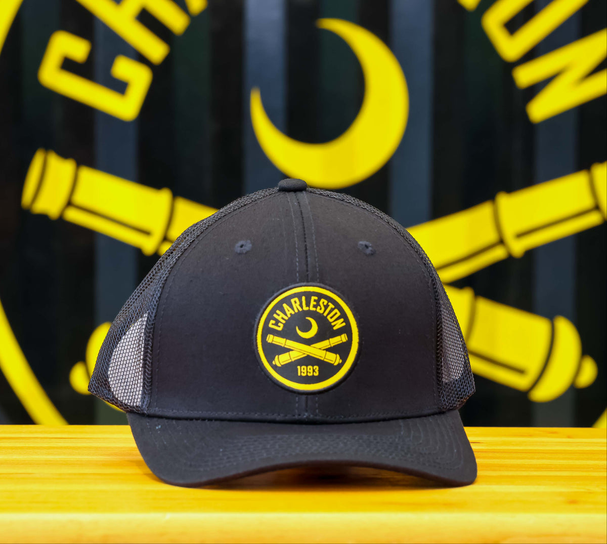 A black trucker-style baseball cap with a yellow circular 'Charleston 1863' patch on the front, resting on a light wood table in front of a large black and yellow Charleston sign.